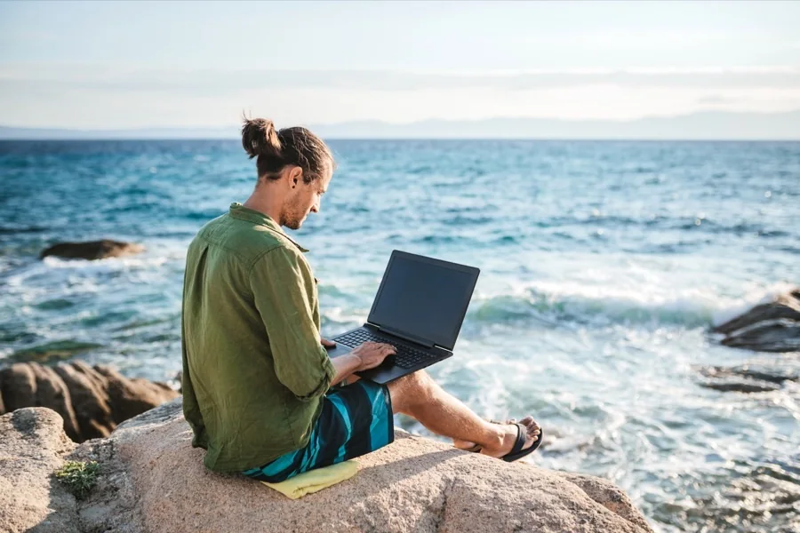 An American working on a laptop from a rocky coastline overseas.